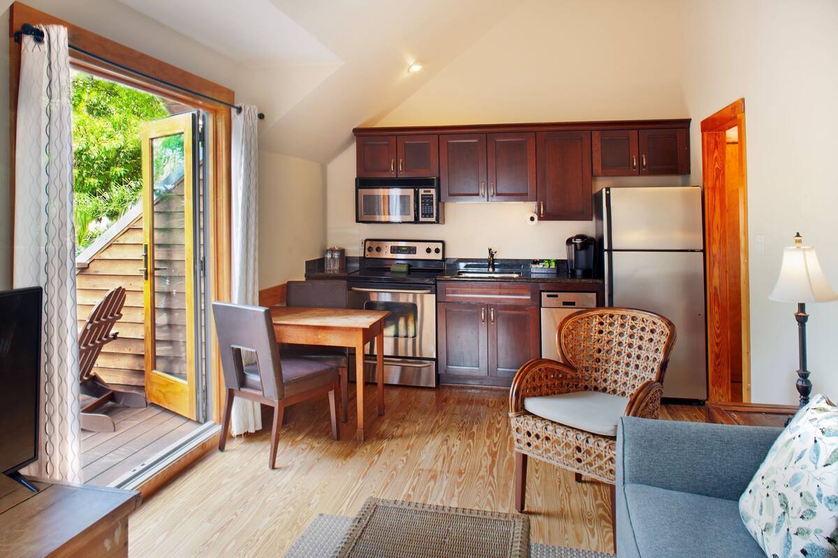 A compact kitchen with wooden cabinets, a microwave, stove, and refrigerator. There is a dining area with a table and chairs, and a sofa.