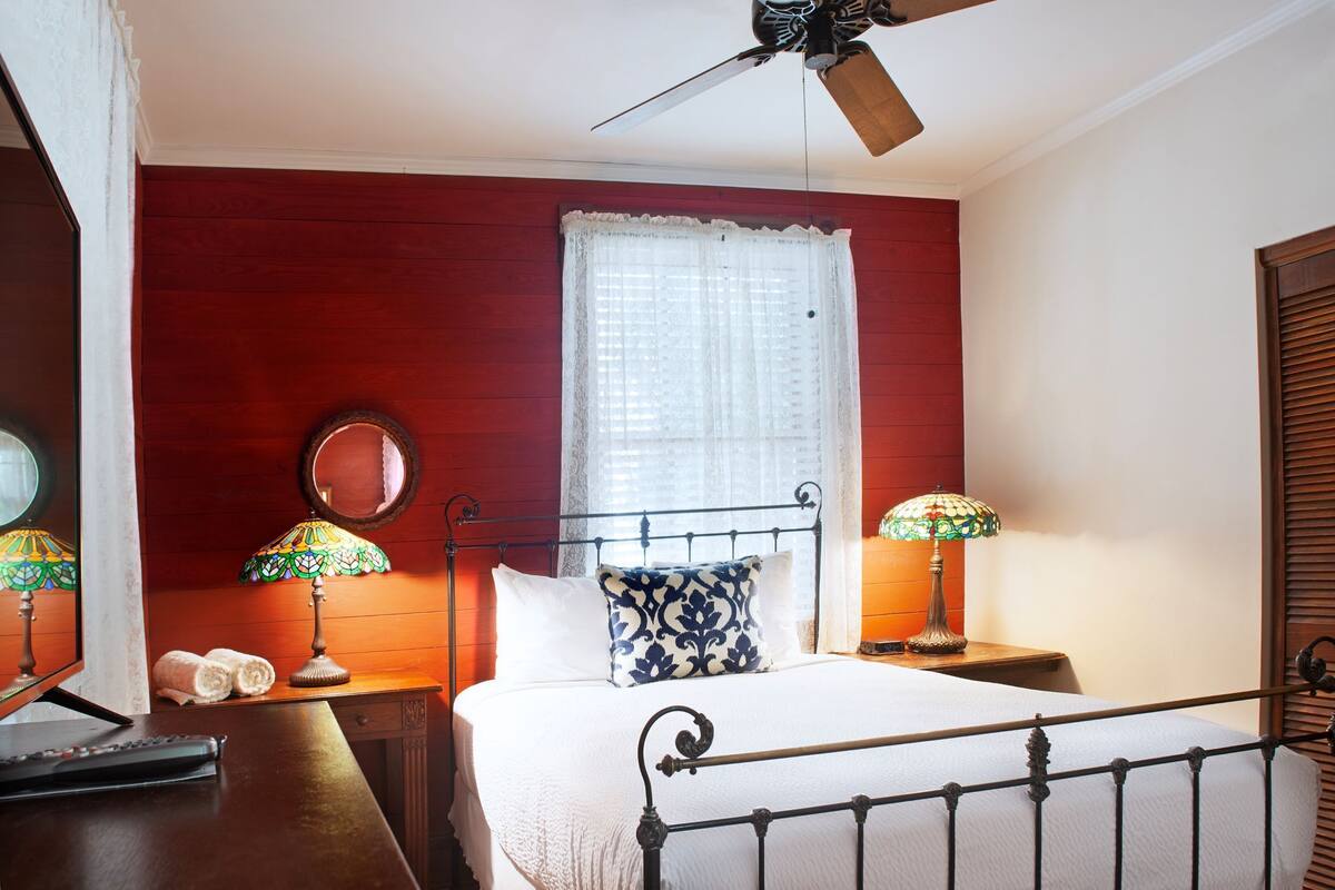 A room with a bed, two bedside lamps, a ceiling fan, a window with lace curtains, and a wooden headboard.
