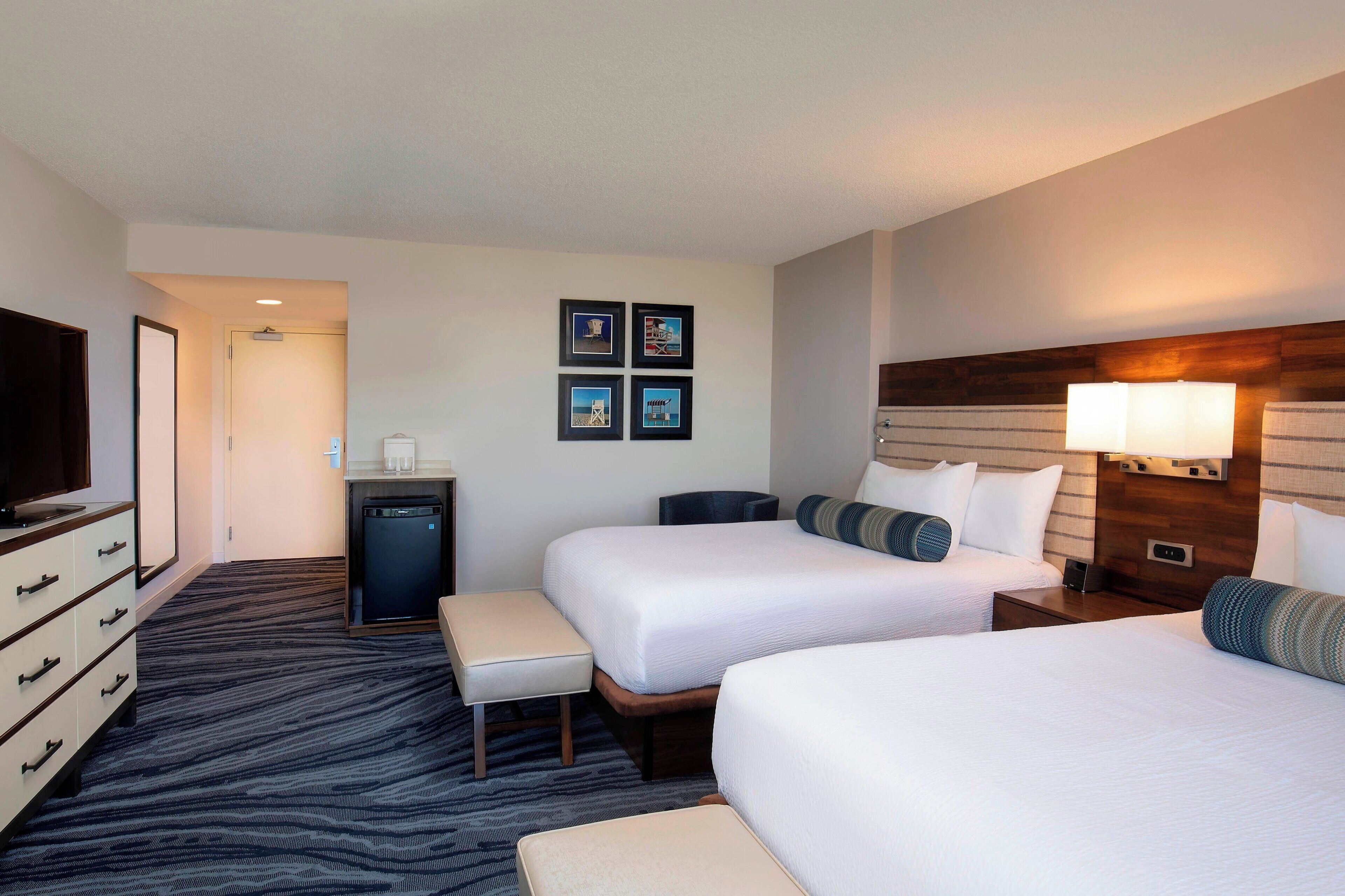 A hotel room with two beds, a desk, a television, and a small kitchenette.