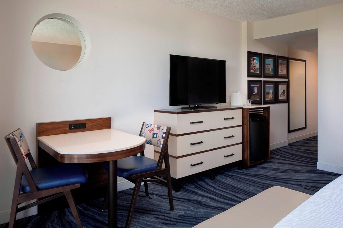 A hotel room with a round mirror, a flat-screen TV, a desk with two chairs, a dresser, and framed pictures on the wall.