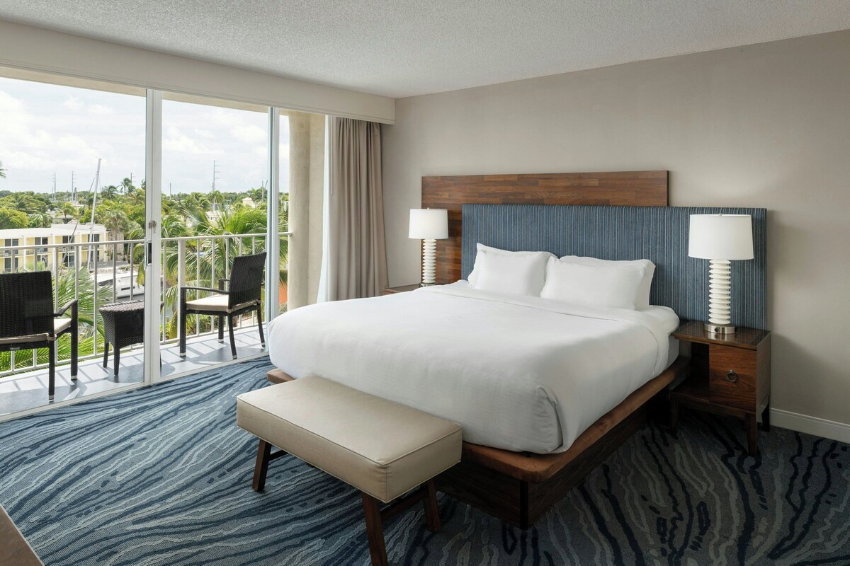 A hotel room with a large bed, a bench, two bedside tables with lamps, a balcony with chairs, and a view of palm trees and buildings.