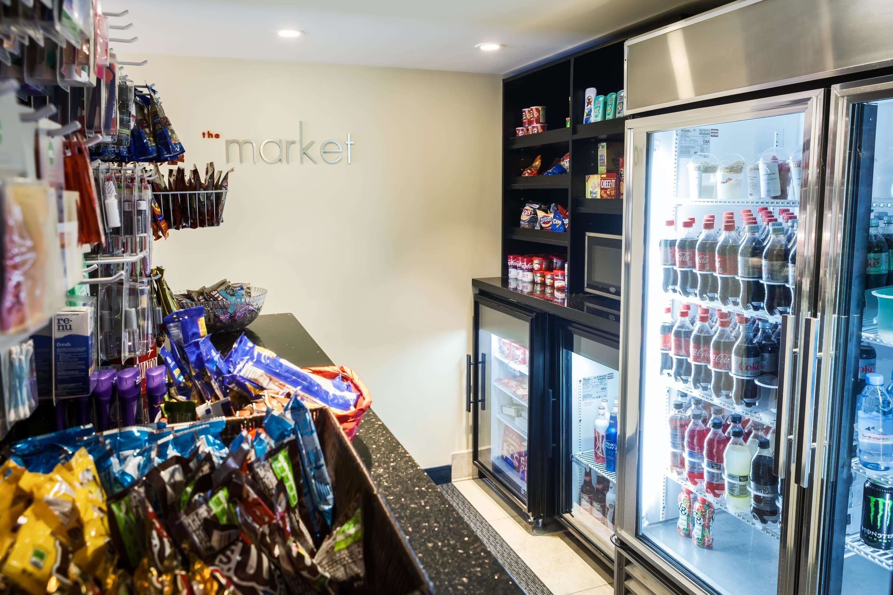 Un expositor refrigerado con diversas bebidas, un cartel en la pared que dice "the market" y estantes llenos de aperitivos y productos envasados.