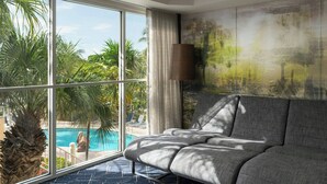 Una habitación moderna con un ventanal grande, un sofá gris, una lámpara y vistas a una piscina y palmeras.