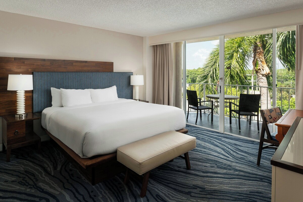 A hotel room with a large bed, a bench, a desk, and a balcony with chairs and a table.
