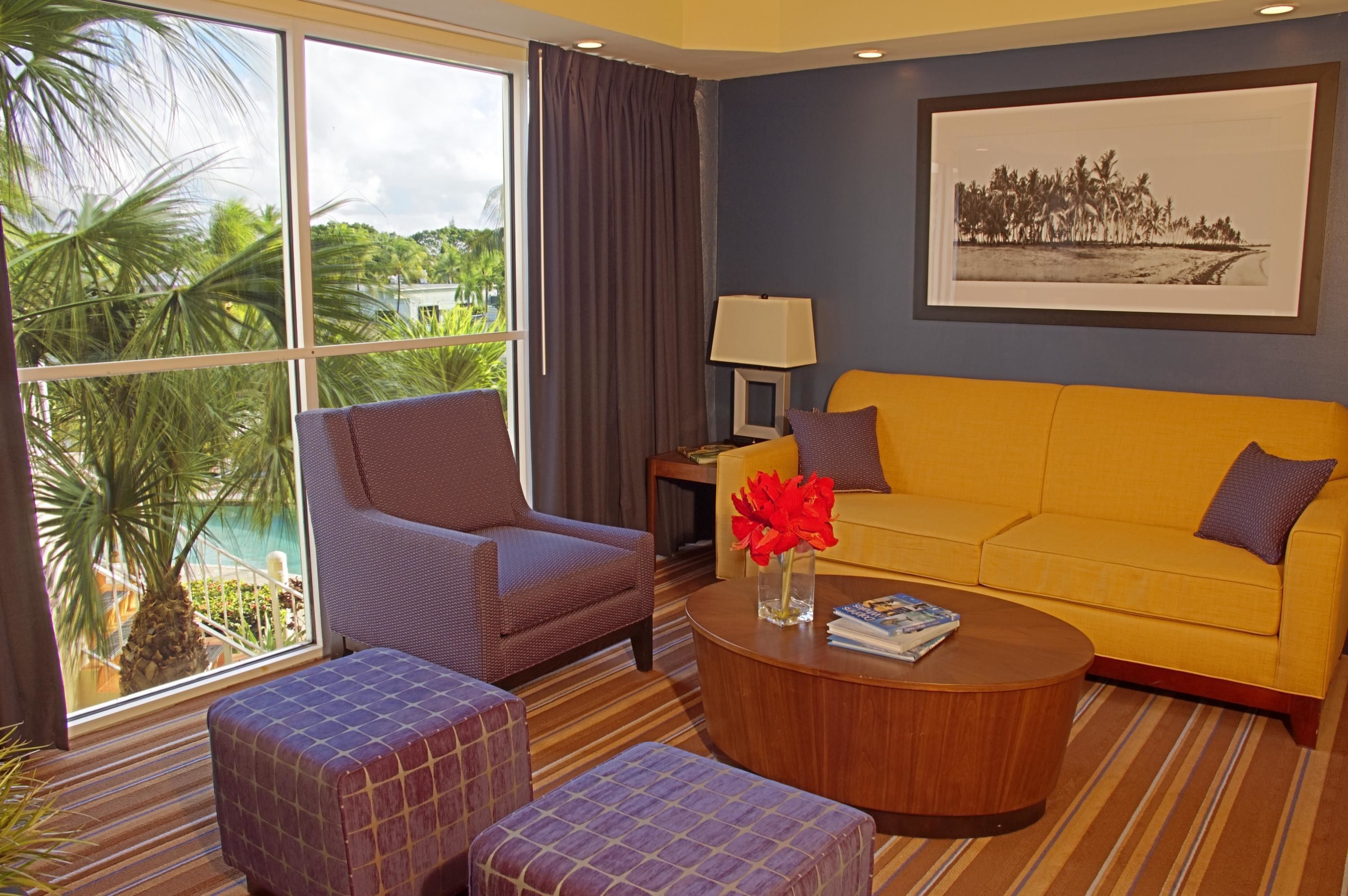 A room with a large window, a yellow sofa, a brown armchair, and a small table with a vase of red flowers.