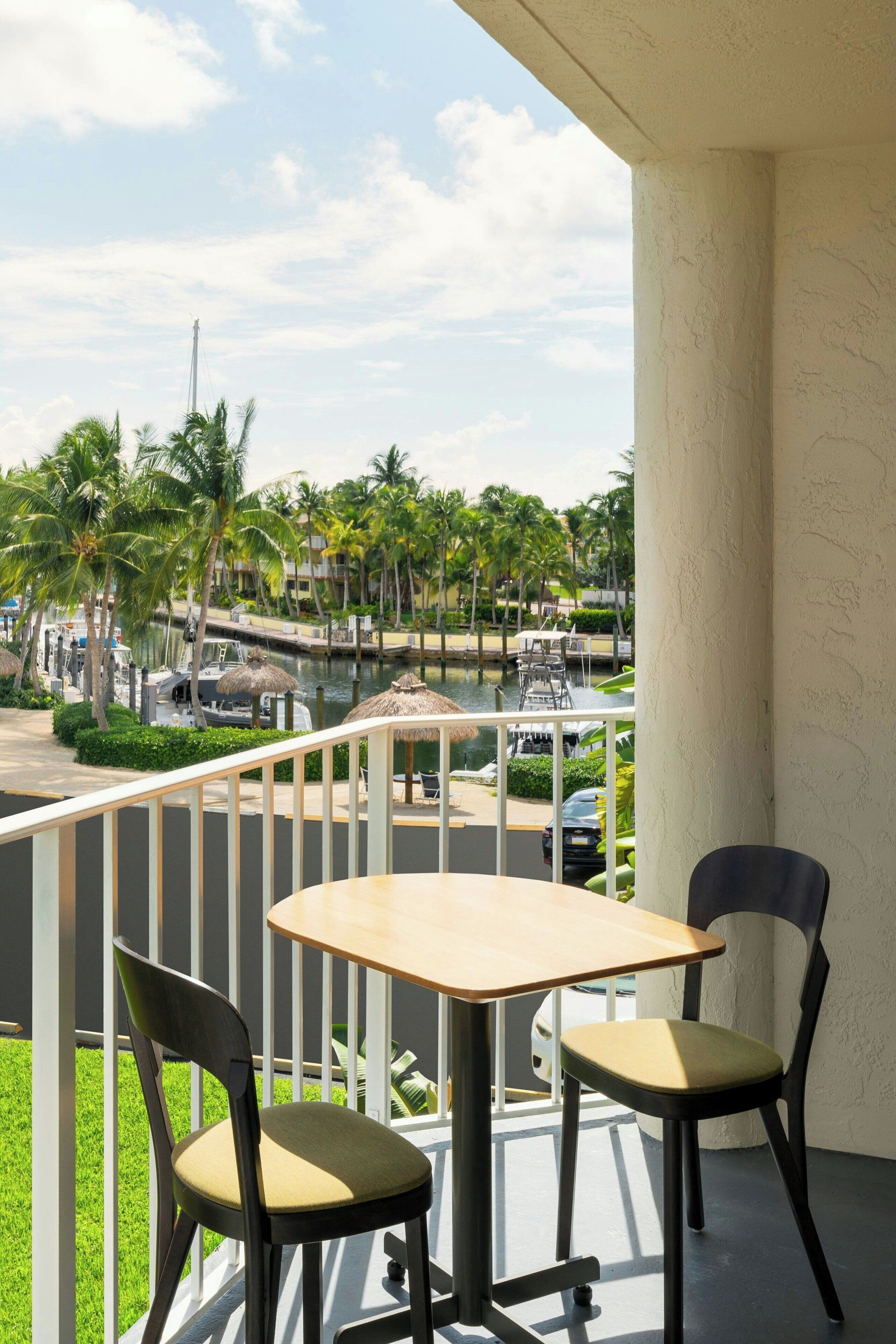 A balcony with a table and two chairs, overlooking a marina with boats and palm trees.