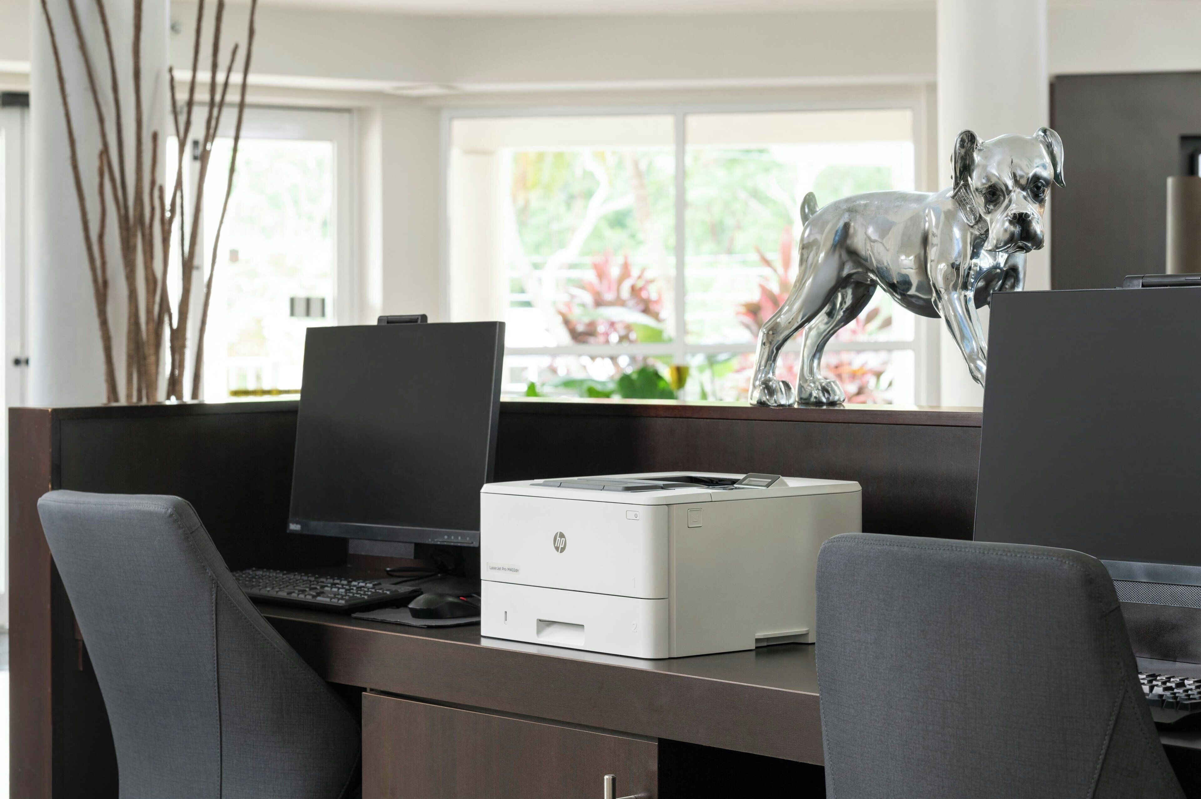 Ein moderner Schreibtisch mit einem Drucker, zwei Monitoren und einer dekorativen Hundefigur.