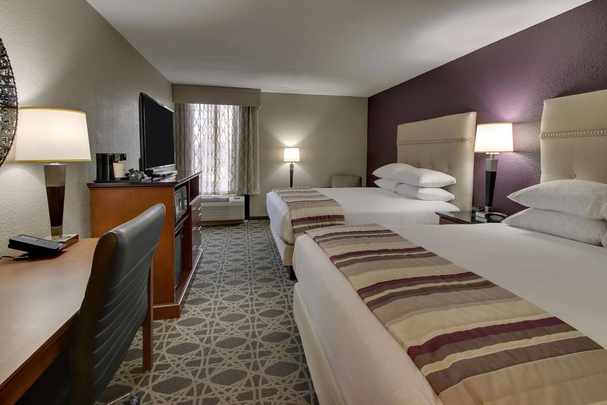 A hotel room with two beds, a desk, a chair, a TV, and a lamp.
