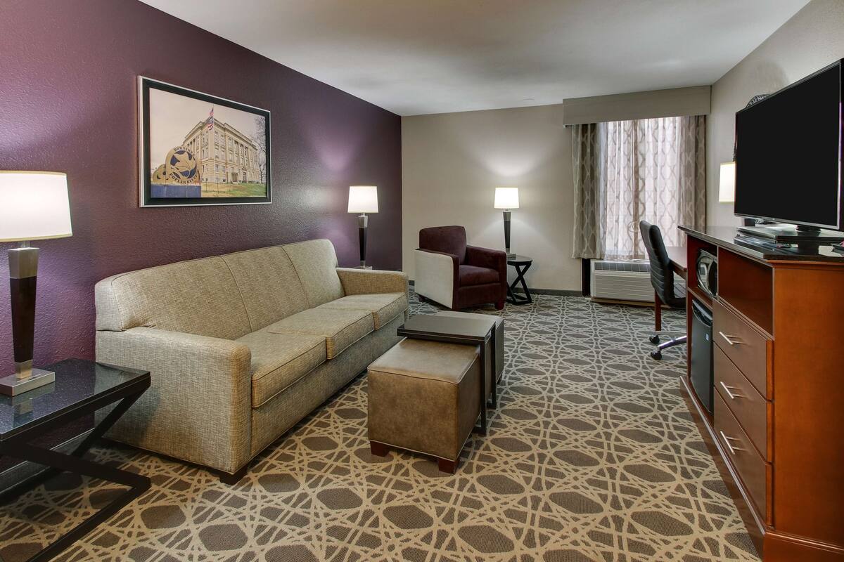 A hotel room with a sofa, a small table, a chair, a TV, and a patterned carpet.