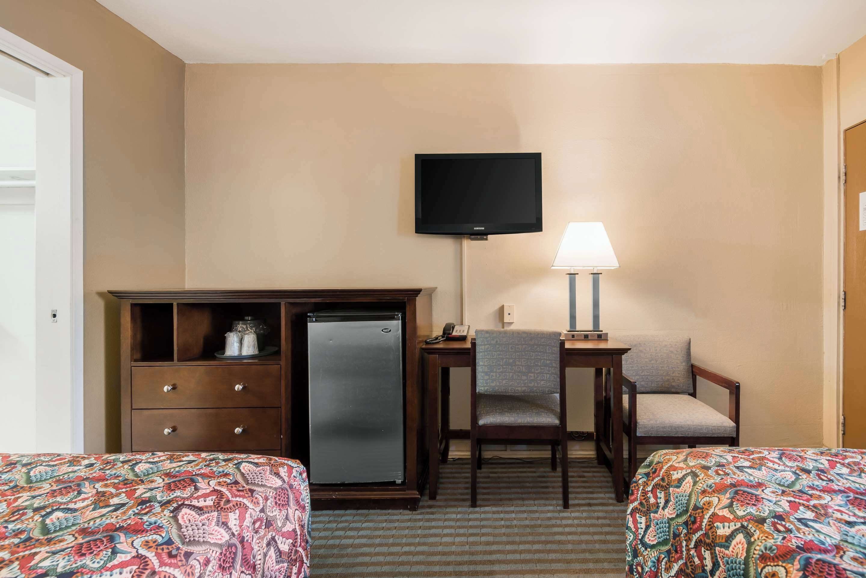 A hotel room with two beds, a desk with a chair, a refrigerator, a television mounted on the wall, and a lamp on the desk.