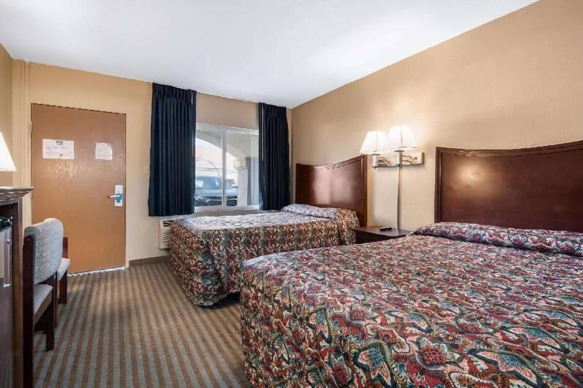 A hotel room with two beds, a desk, a chair, a window with curtains, and a lamp.