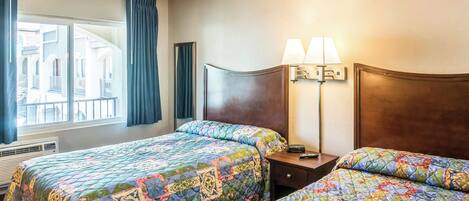 A hotel room with two beds, a large window with curtains, a nightstand with lamps, and a view of buildings outside.