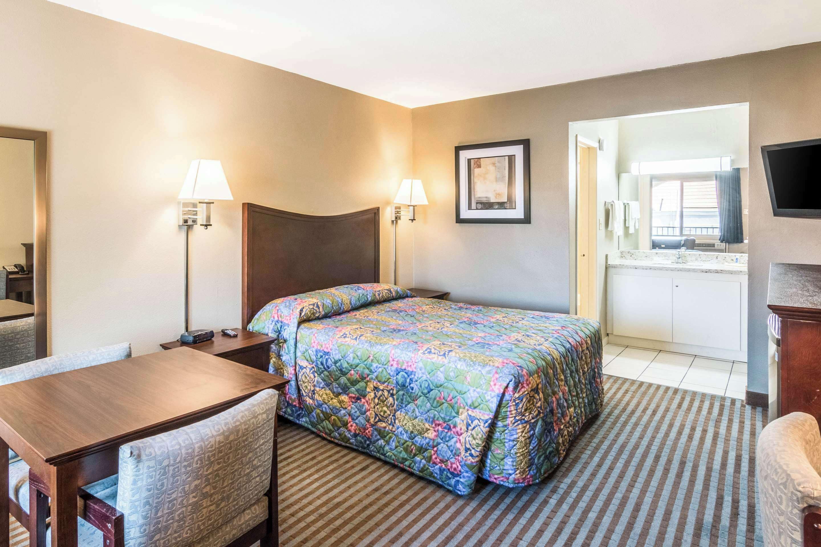 Une chambre d’hôtel avec un lit, un bureau, deux chaises, une télévision et une salle de bain visible à travers une porte ouverte.