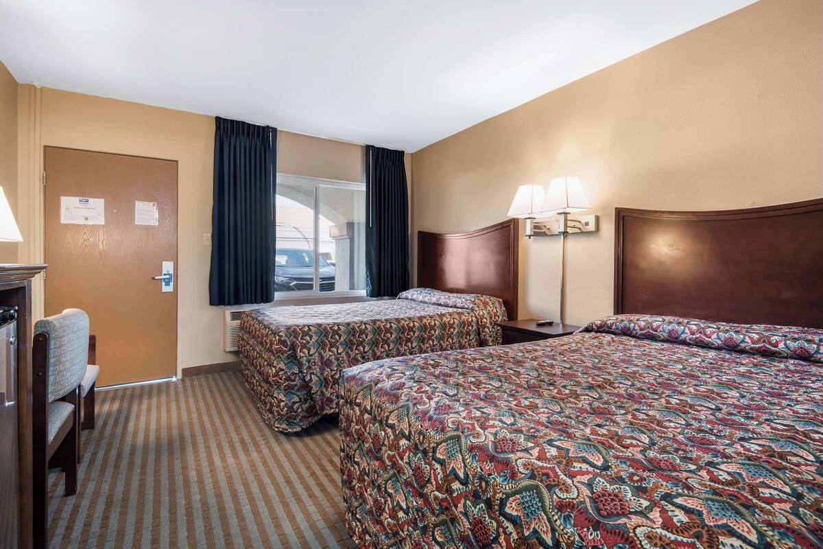 A hotel room with two beds, a desk, a chair, a window with curtains, and a lamp.