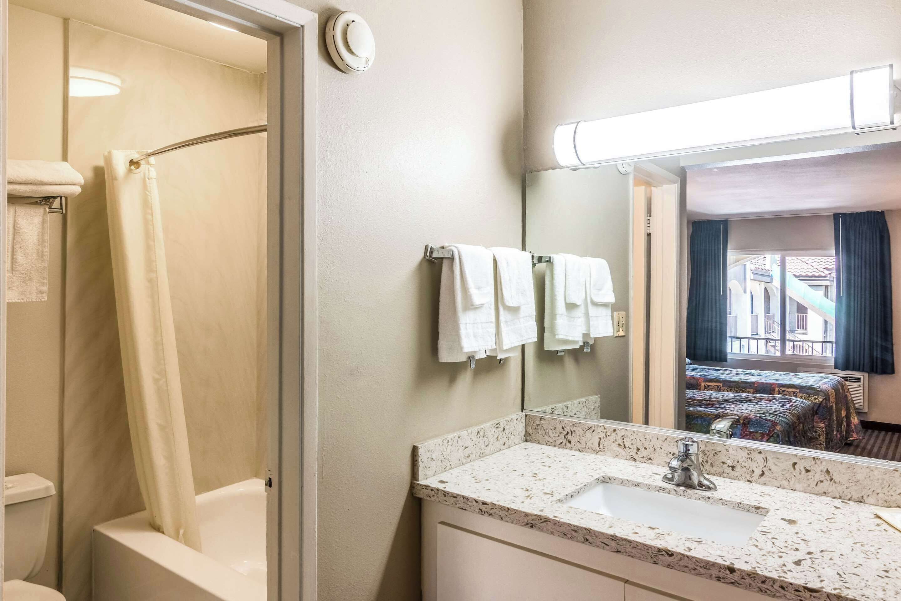 A bathroom with a shower, a sink, and a mirror. There are towels hanging on the wall and a window with curtains.