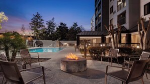 An outdoor pool area with a fire pit and seating, surrounded by trees and a building.