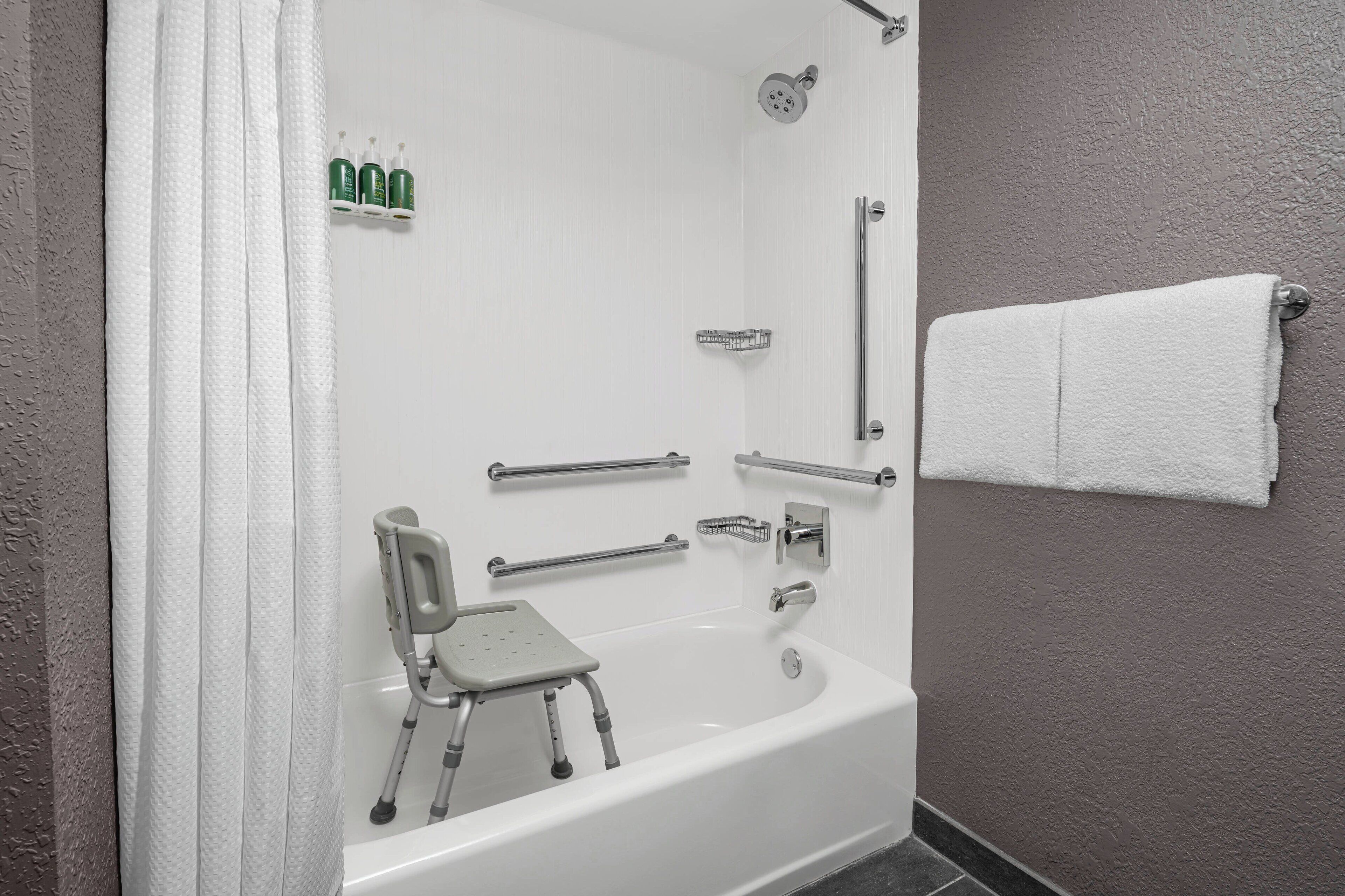 Un baño con bañera blanca, barras de apoyo, cabezal de ducha y un perchero para toallas.