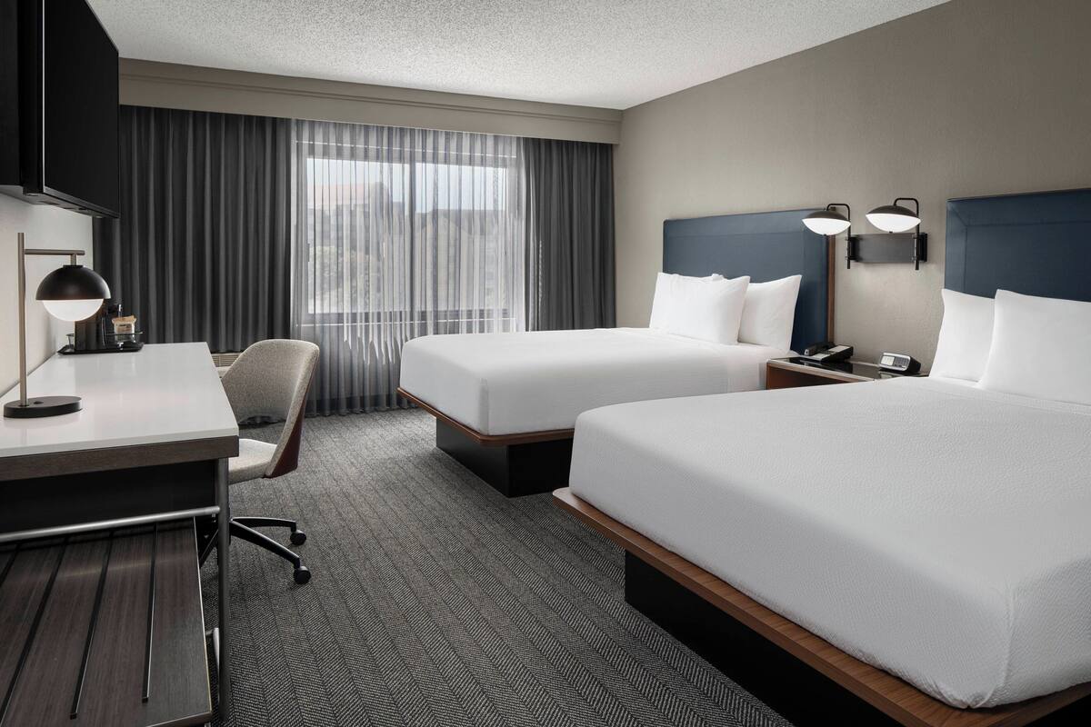 Een hotelkamer met twee bedden, een bureau met lamp, een stoel en een groot raam met gordijnen.