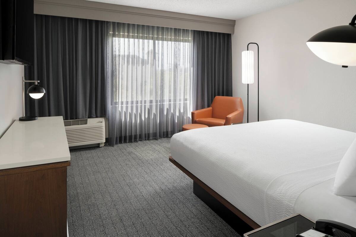 Een hotelkamer met een groot bed, een bureau met een lamp, een stoel en een raam met gordijnen.