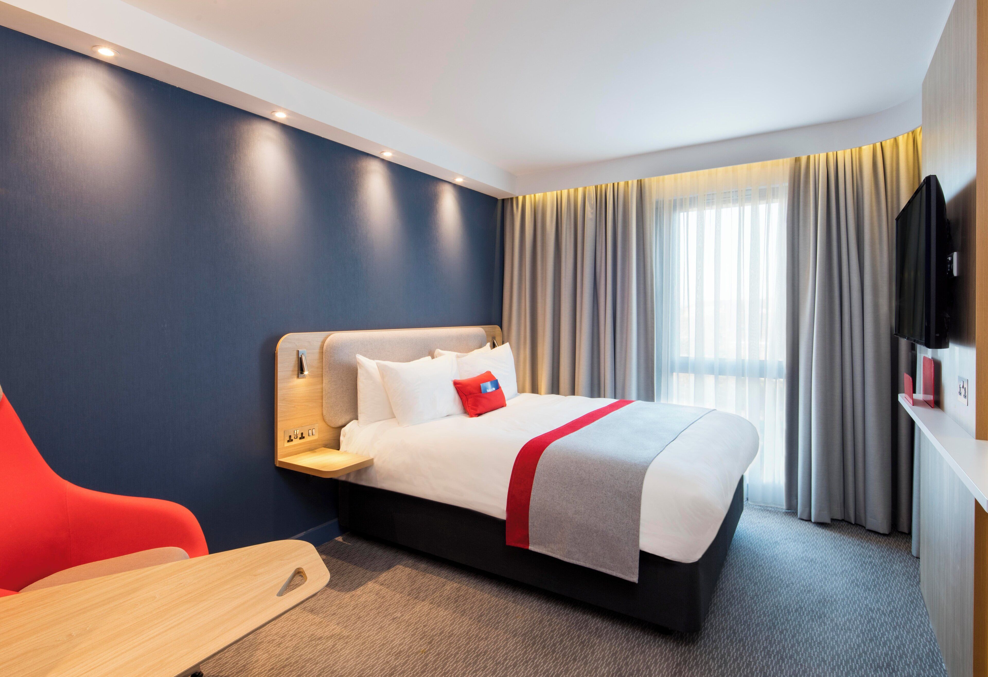 A hotel room with a bed, a television, a red chair, and a wooden side table.