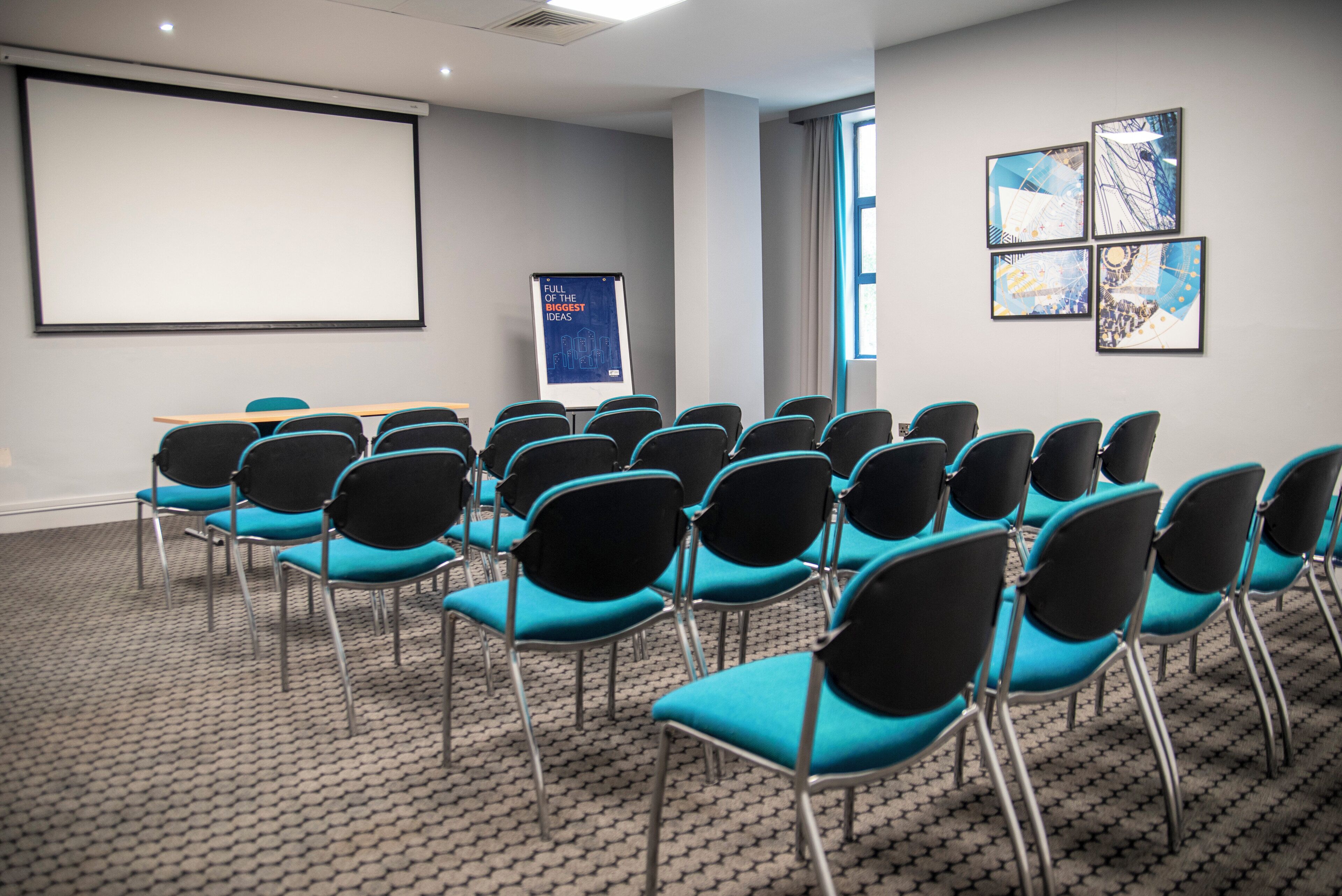 A conference room with rows of chairs, a projector screen, a presentation board, and framed artwork on the wall.