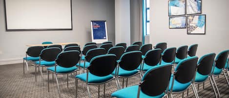 A conference room with rows of chairs, a projector screen, a presentation board, and framed artwork on the wall.