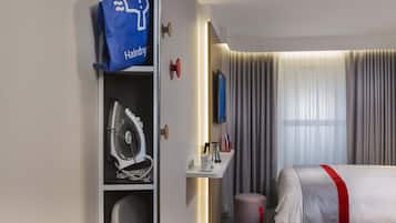A hotel room with a bed, a desk with a lamp, a hairdryer bag, and a TV.