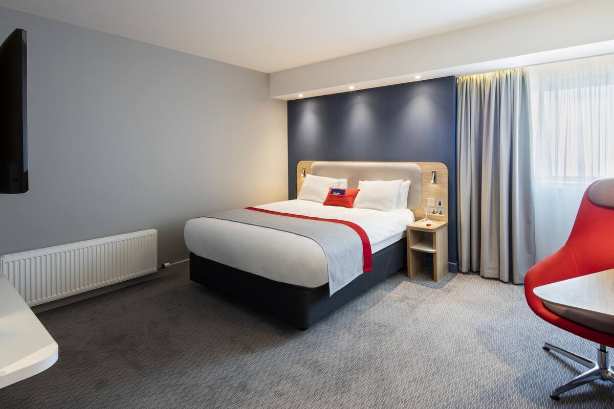 A modern hotel room with a large bed, a bedside table, a red chair, and a view of the outdoors through a window.