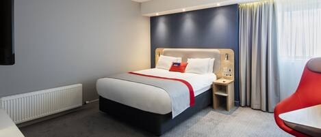 A modern hotel room with a large bed, a bedside table, a red chair, and a view of the outdoors through a window.