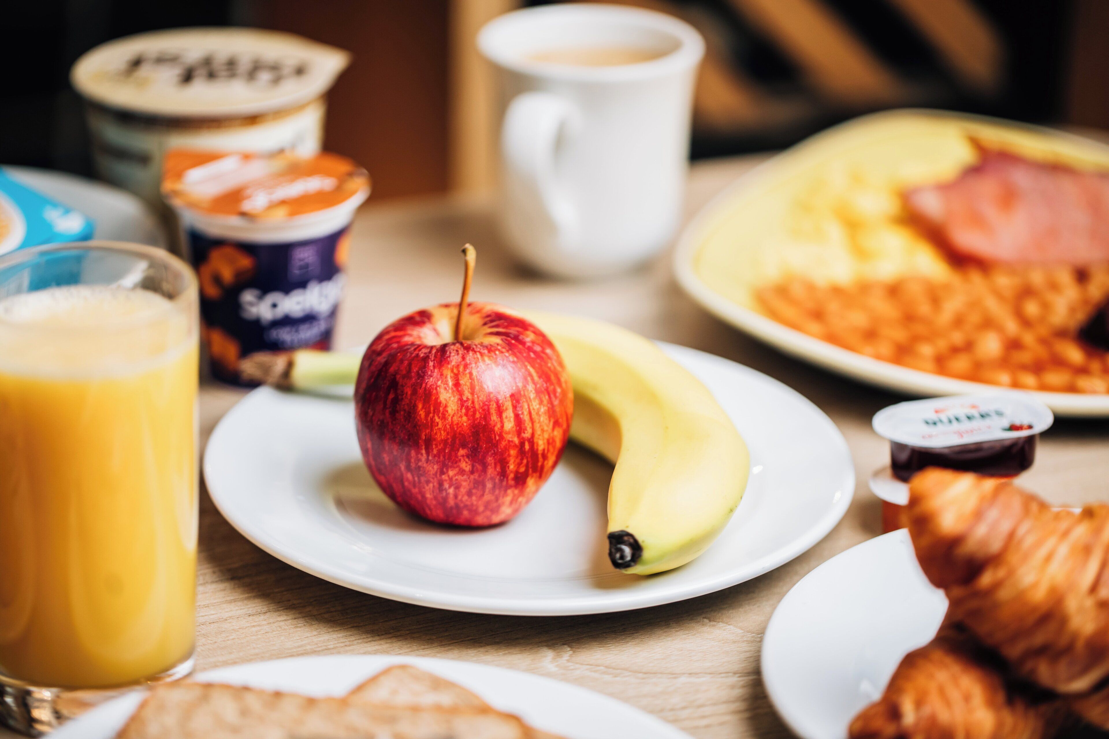A breakfast setting with a plate of eggs, a banana, an apple, a croissant, a glass of orange juice, a cup of coffee, and a container of yogurt.