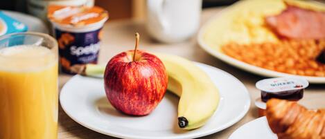 A breakfast setting with a plate of eggs, a banana, an apple, a croissant, a glass of orange juice, a cup of coffee, and a container of yogurt.