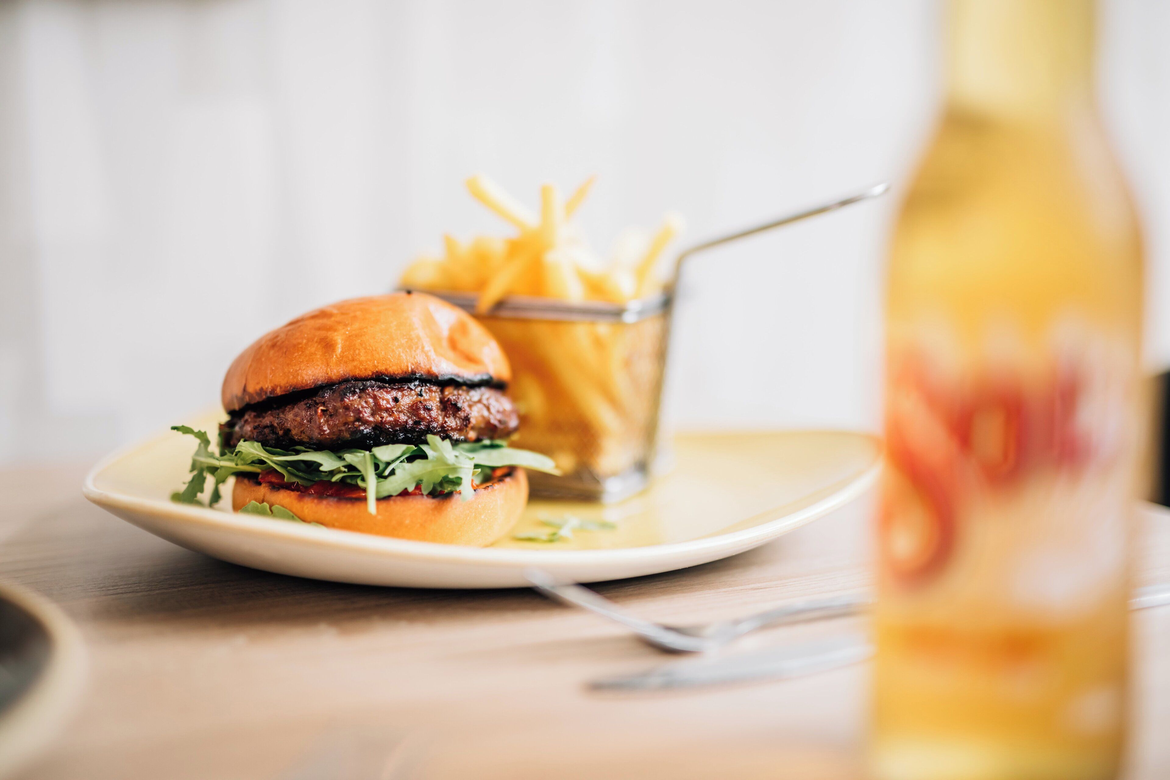 A plate with a burger and fries, a glass of a yellow beverage, and a bottle on a table.