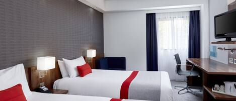 Habitación de hotel con dos camas, un escritorio, una silla, un televisor y una ventana con cortinas.