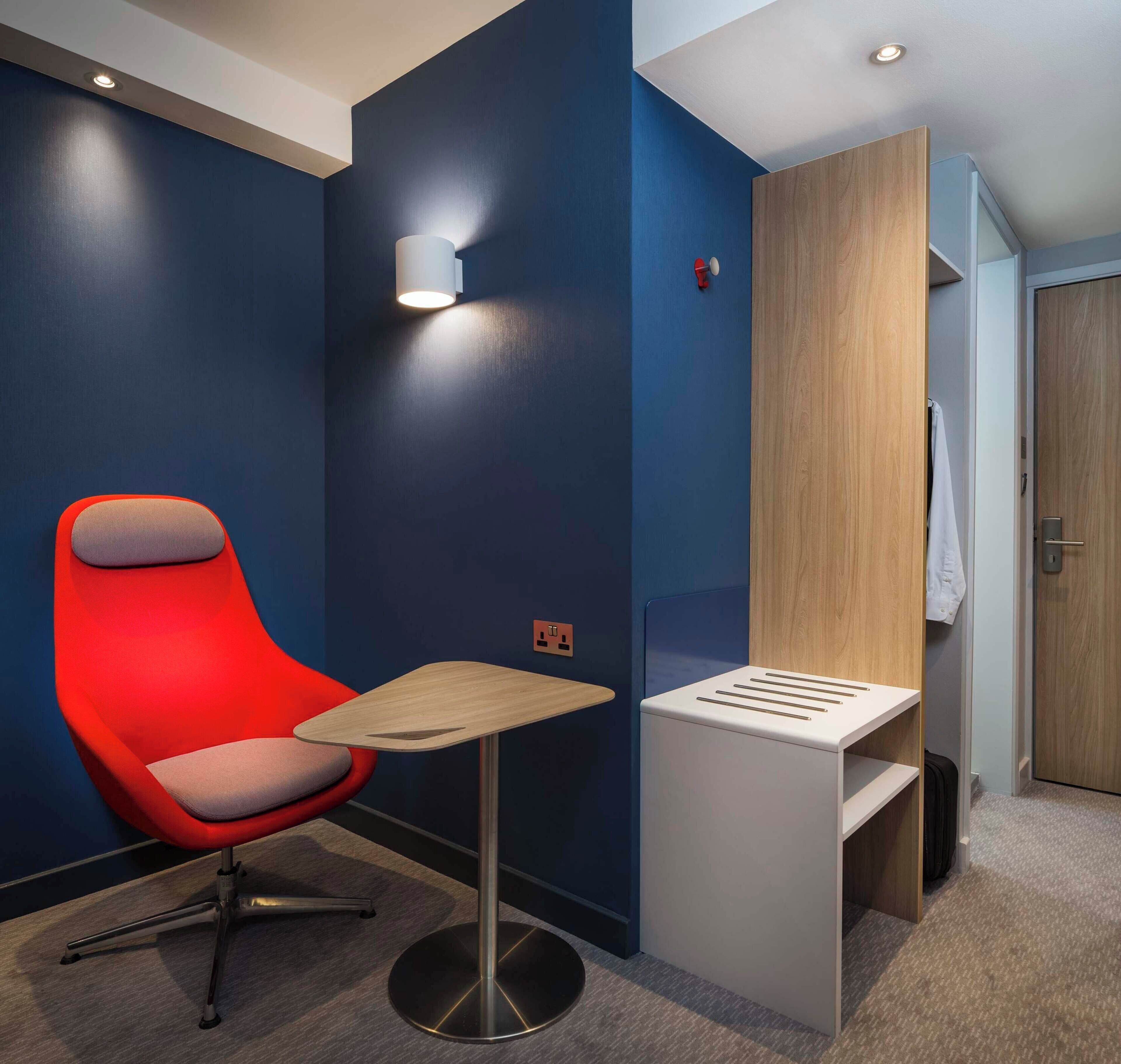 A modern hotel room with a red chair, a small table, a wall-mounted light, and a wooden wardrobe.