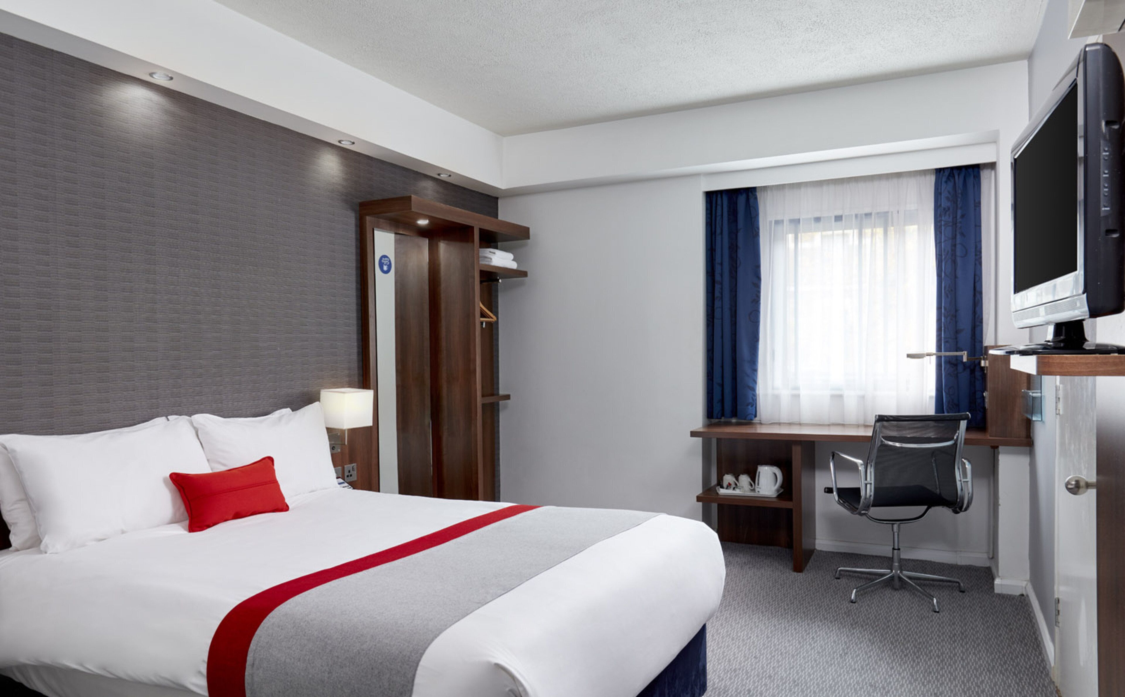 Une chambre d’hôtel avec un lit, un bureau, une chaise, une télévision et une fenêtre avec des rideaux.