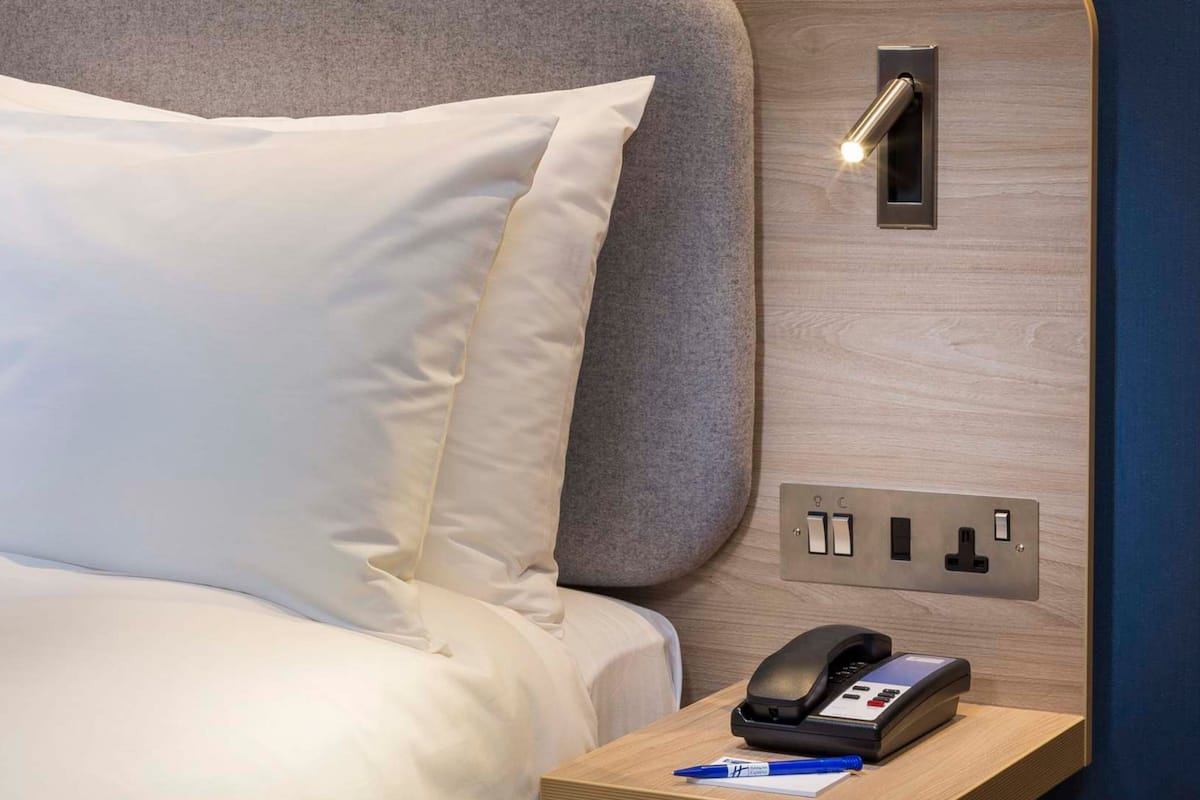 A hotel room with a bed, a bedside table with a phone and a pen, and a wall-mounted light.
