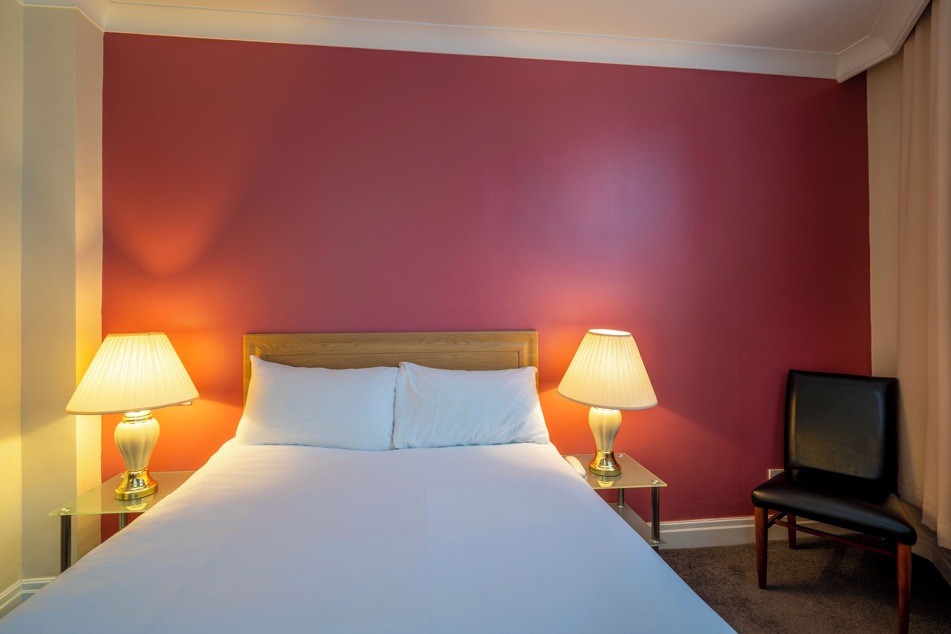 A hotel room with a large bed, two bedside lamps, a wooden headboard, and a black chair.