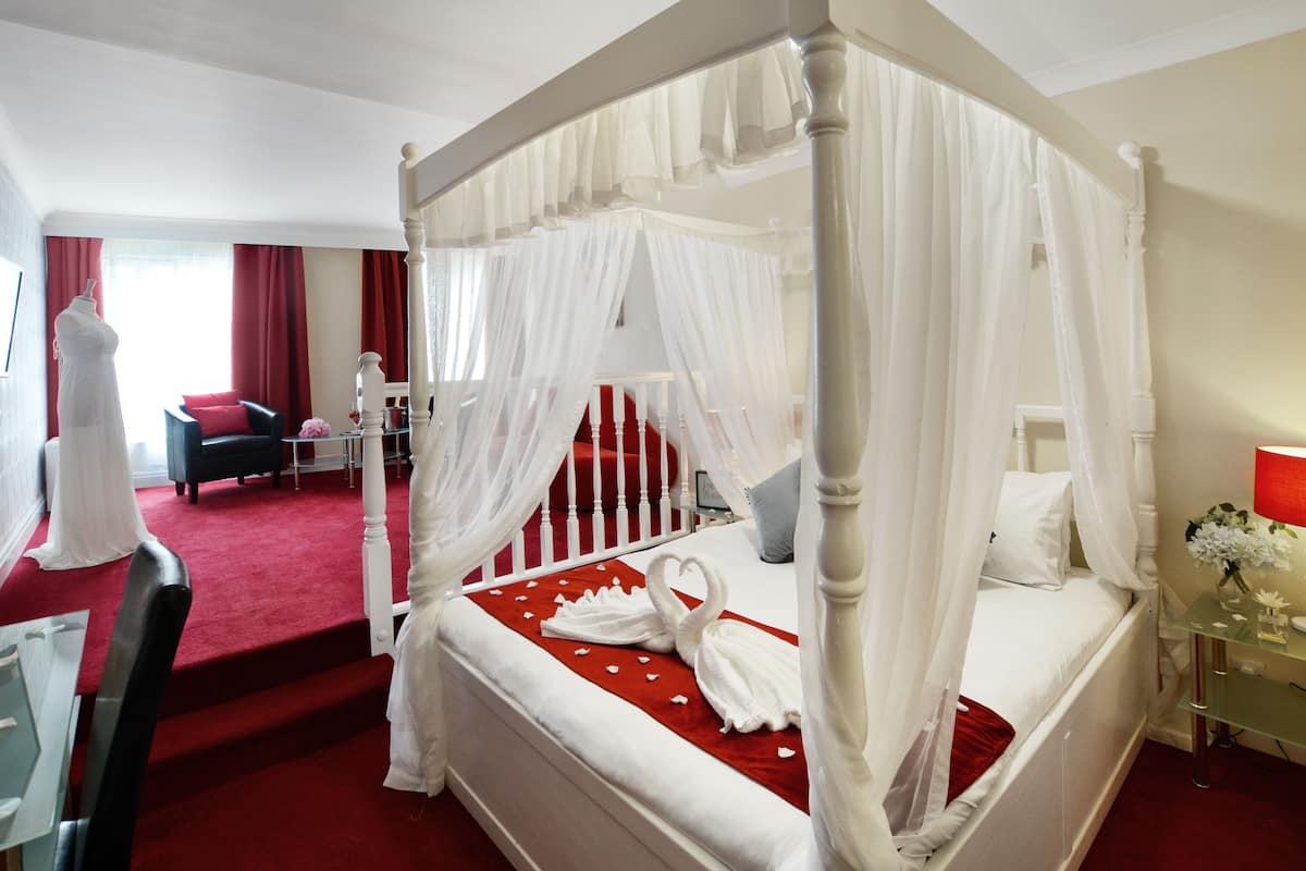 A four-poster bed with a canopy, a red carpeted floor, a small bedside table with a vase of flowers, and a chair in the background.