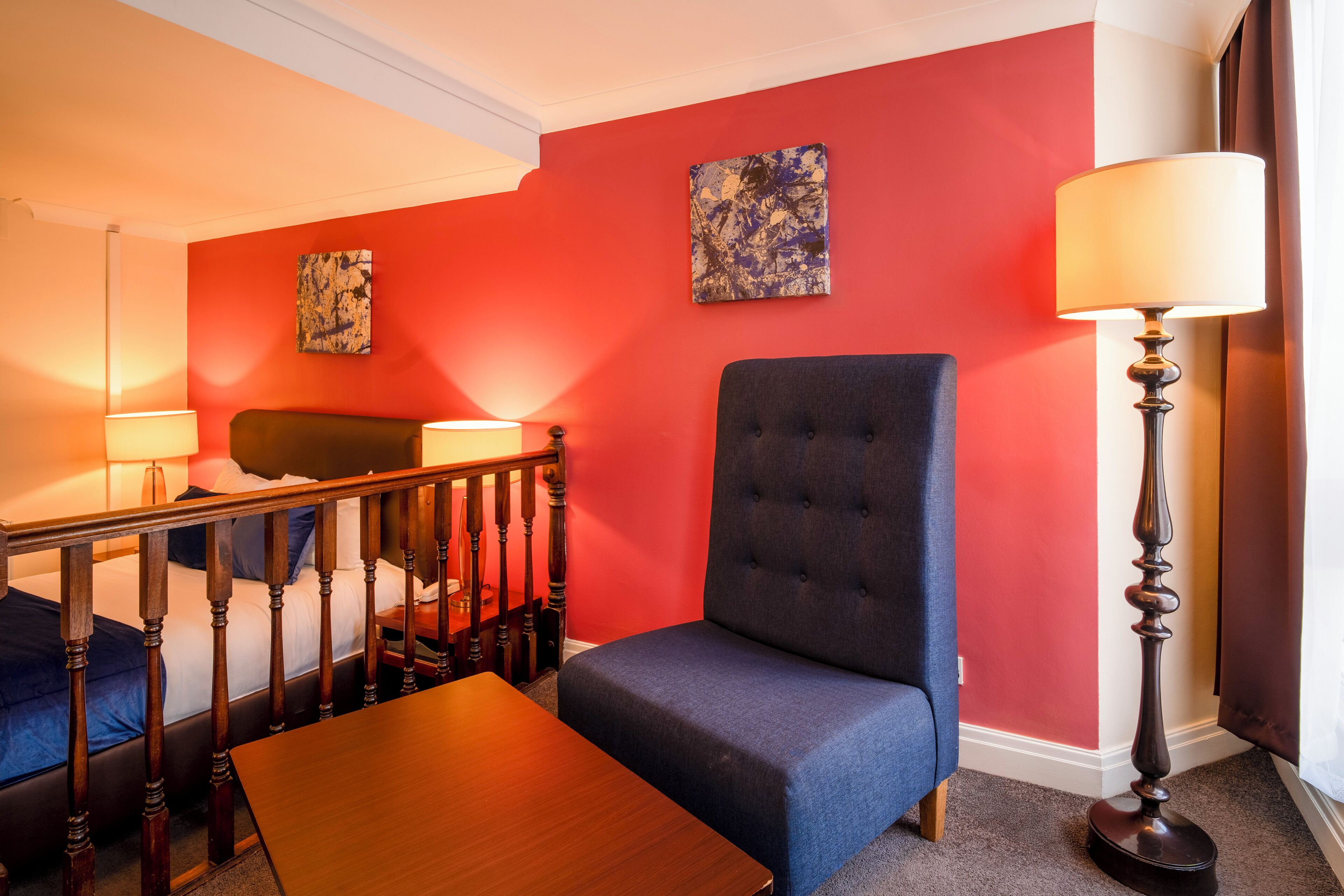 A hotel room with a red wall, a blue sofa, a wooden table, and two lamps.