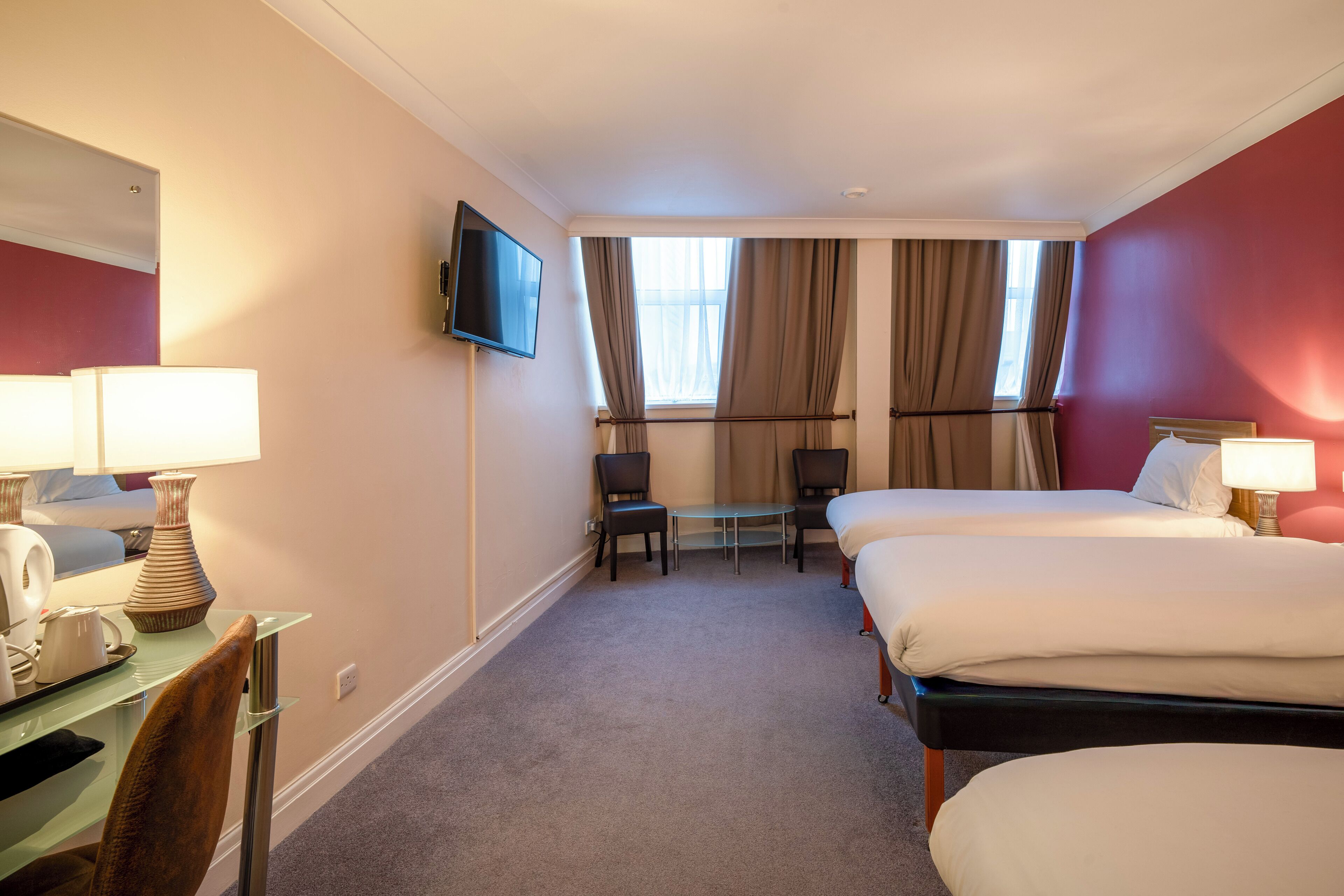 A hotel room with two beds, a desk with a lamp, a chair, and a TV on the wall.