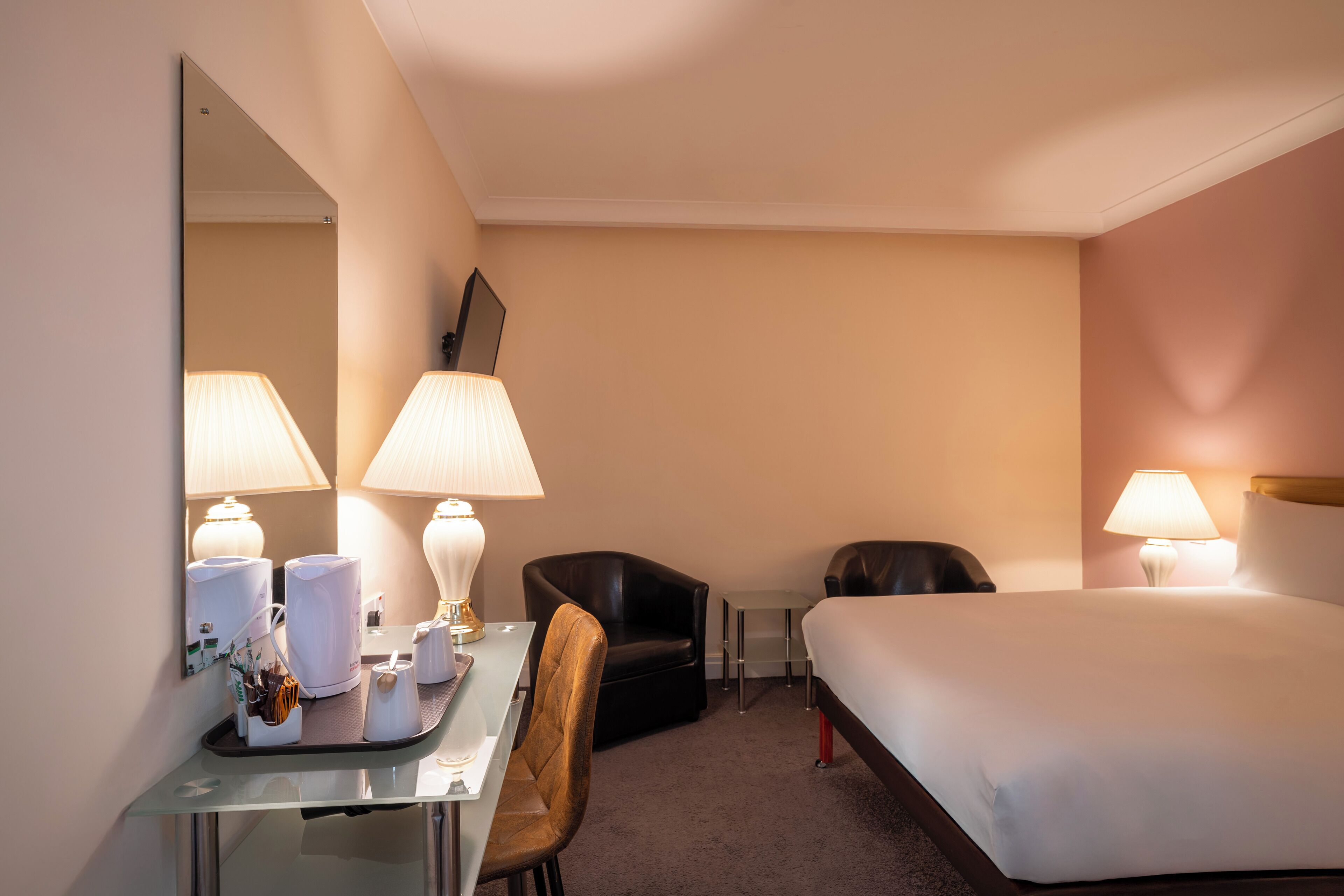 A hotel room with a bed, two chairs, a desk with a lamp, a mirror, and a tray with toiletries.