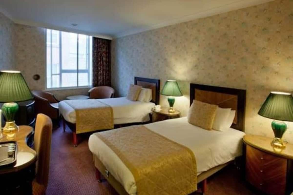 A hotel room with two beds, a desk, chairs, lamps, and a window.