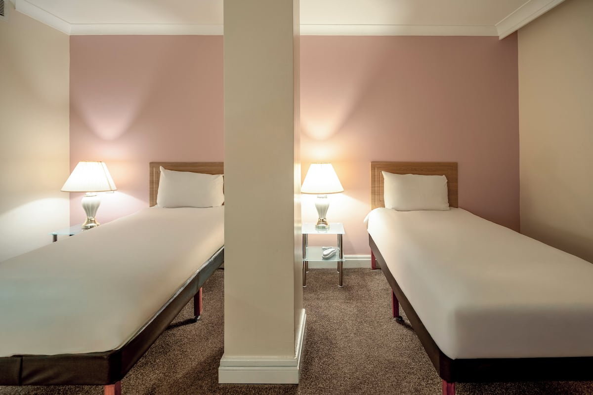 A hotel room with two single beds, each with a bedside table and lamp, and a carpeted floor.