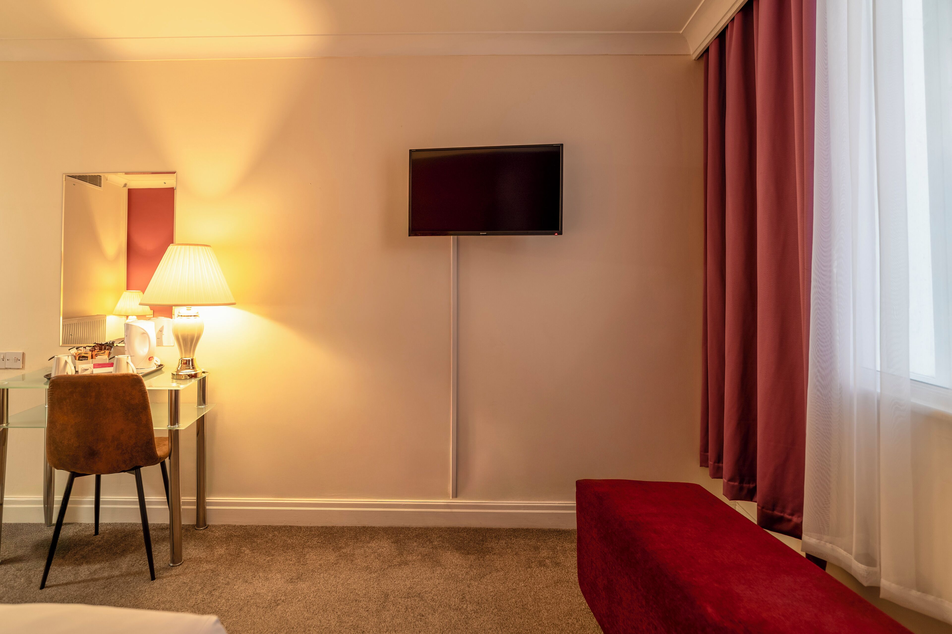Quarto de hotel com televisão de tela plana na parede, escrivaninha com luminária, cadeira e um banquinho vermelho.