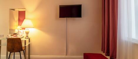 Quarto de hotel com televisão de tela plana na parede, escrivaninha com luminária, cadeira e um banquinho vermelho.