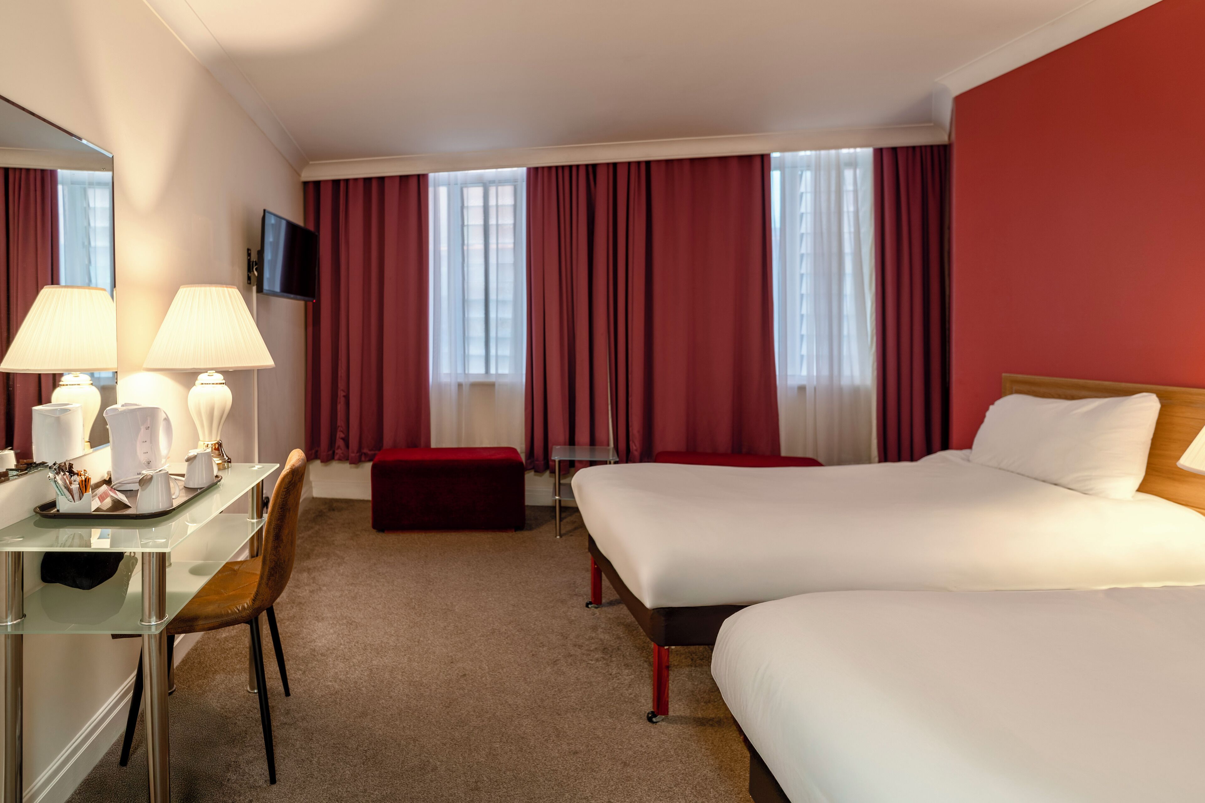 A hotel room with two beds, a desk with a chair, a TV mounted on the wall, and a window with red curtains.