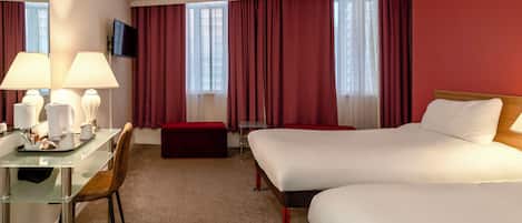 A hotel room with two beds, a desk with a chair, a TV mounted on the wall, and a window with red curtains.