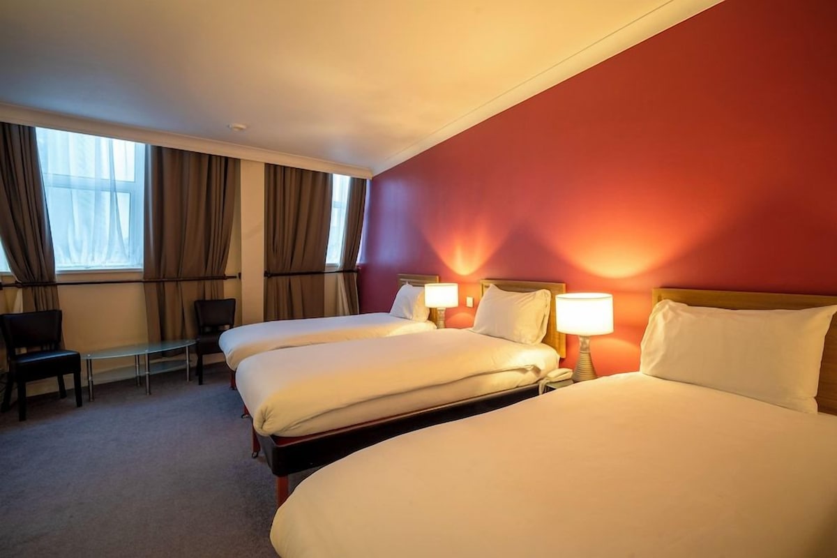 A hotel room with two beds, a desk, chairs, and a window with curtains.