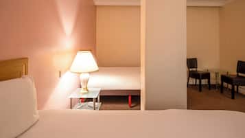 A hotel room with a bed, a bedside table with a lamp, a chair, and a small table with chairs in the background.