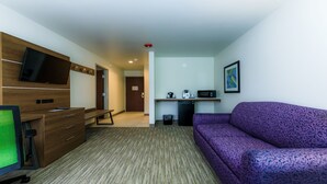 Hotelkamer met een houten bureau, een flatscreen televisie, een paarse bank, een kitchenette met een magnetron en een prullenbak.